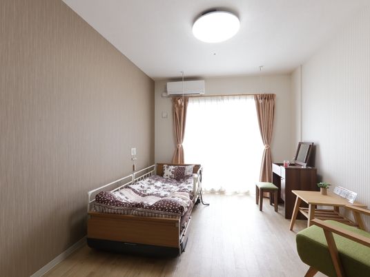正面は大きなガラス張り、焦げ茶色の壁際には介護用ベッドが置かれている。白グレイのストライプの壁際にはドレッサーなどが置かれている。
