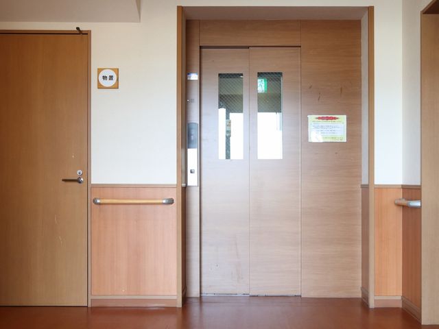 木目調のダブルドア廊下 エレベーター ゆったりとした木目調のドアは、多用途に活用される快適な多目的室へと繋がっています。職員の皆さんが利用者様の快適な生活をサポートするための、大切なスペースとなっています。