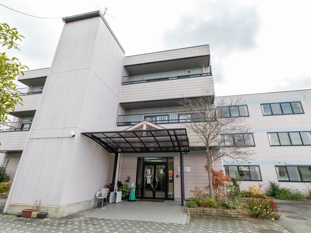 白壁の建物とエントランス 外観 手入れが行き届いた緑に囲まれた明るいエントランスです。どなたでも心地よくご来訪いただけるように配慮されています。