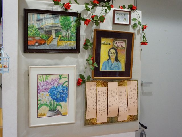 花と絵画の飾られた壁 壁の展示 壁面には彩り豊かな花と絵画が飾られ、心温まる雰囲気を醸し出しています。手書きの文字が添えられた掲示物も見受けられます。