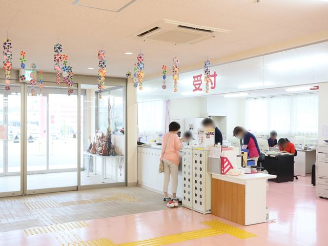 エントランスの賑やかな様子 玄関 彩り豊かな飾りが天井に吊るされた受付エリアで、スタッフが来訪者の対応をしています。手前にはバリアフリーのための斜めになった通路が見えます。