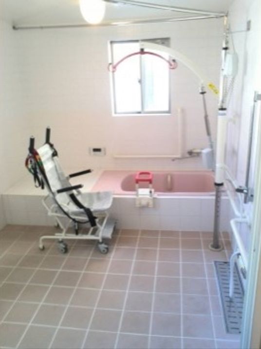 淡い色合いの明るい浴室。浴槽や壁回りなど各所に手すりがついている。車椅子型のシャワーチェアが置かれ、浴室内を座ったまま移動できる。