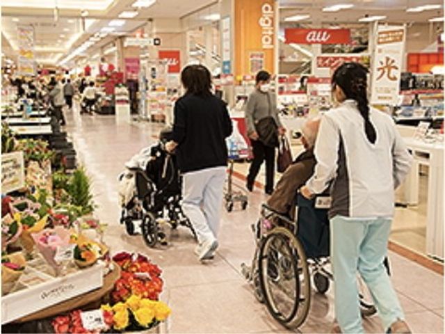 車椅子利用者とスタッフ レクリエーション スタッフが車椅子を押しながらお散歩に出かける様子。日常の光景ですが、ここでの働きがいを感じていただけるでしょう。