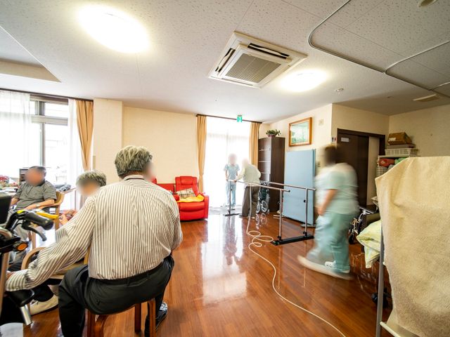 日常の様子が伝わる居室 機能訓練室 柔らかな光が差し込む快適な空間で、利用者が過ごされています。スタッフがたくさん動き回っており、活気が感じられます。