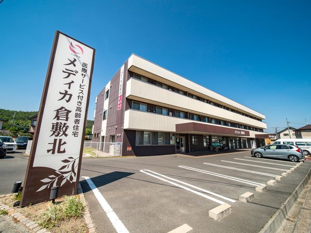 施設の看板と外観 駐車場 清々しい青空の下、落ち着いた色合いの建物が広々とした駐車場を備えています。