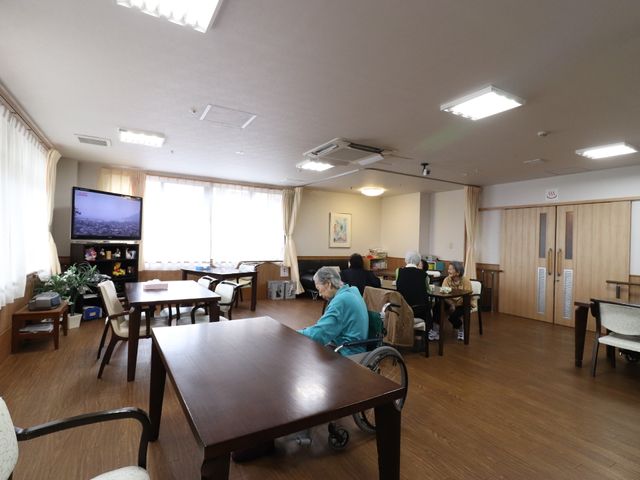 広々とした共有スペース テーブルの高さ ゆったりとしたダイニングで利用者が食事を楽しんでいます。大画面テレビがあり、リラックスした空間を提供しています。