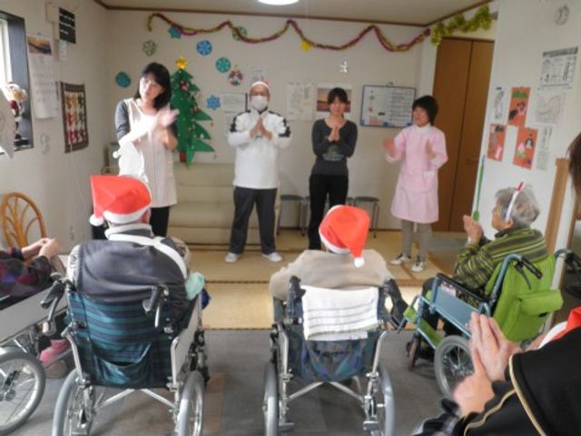多目的室でのイベント活動 クリスマス会 サンタ帽をかぶった利用者が集い、スタッフと共に手拍子をしている賑やかな様子。明るく広々とした空間でイベントを楽しむ姿が印象的です。