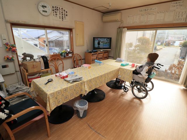 共用スペースの日常風景 食堂 明るく開放感のある食堂で、利用者の方が日差しを浴びながら静かに時間を過ごされています。テーブルには共有の物品が整然と並び、温かなコミュニティの一端を垣間見ることができます。