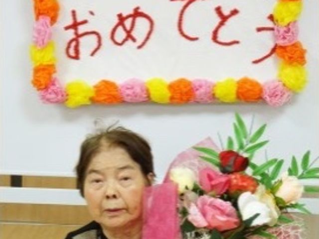 お祝いの花と笑顔の方 お誕生日会 温かみのある掲示に囲まれ、花束をお手元に笑顔を見せる方が、ここでの穏やかな時間を物語っています。