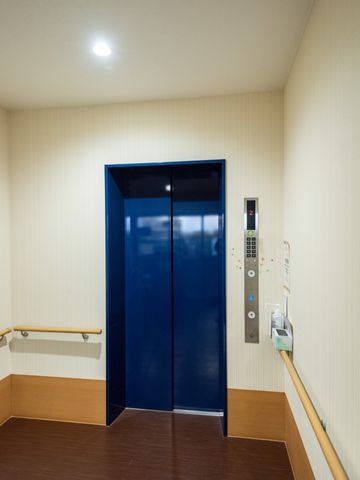 青い扉のエレベーター エレベーター 手すり付きのホールにある青い扉のエレベーターは、バリアフリー設計です。利便性を考慮した明るく落ち着いた色合いが特徴です。