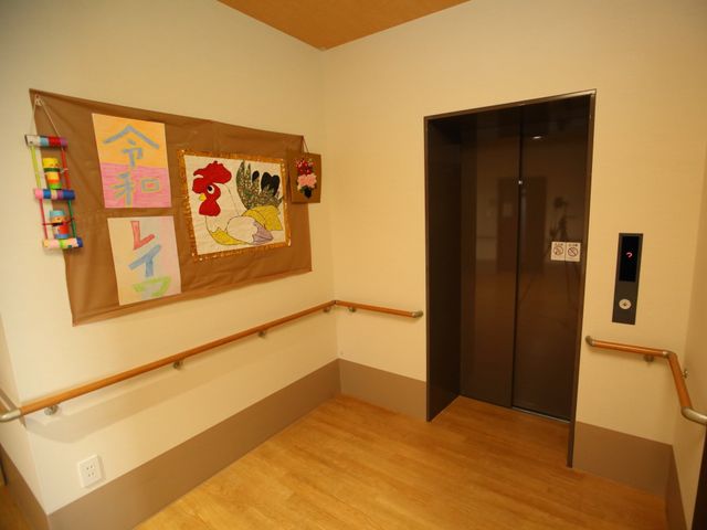 廊下の壁飾りとエレベーター 廊下 手作りの壁飾りが暖かみのある廊下に彩りを与えています。ゆったりとしたスペースで誰もが快適に行き交える設計です。