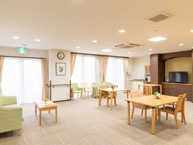 共有スペースの明るい居室 共有スペース ゆったりとした椅子と木のテーブルが配置され、テレビが備えられた快適なラウンジです。窓からの自然光が温かみを感じさせ、居心地の良い空間を提供しています。