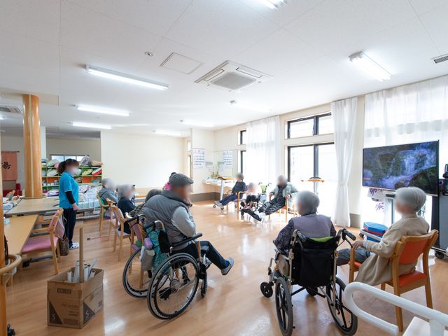 日中の活動スペース レクリエーション 明るく開放的な多目的室で、利用者の方々がテレビを見ながら憩いの時間を過ごされています。車椅子の方もゆとりを持ってくつろげる配置になっております。