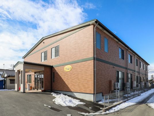 施設は２階建てである。レンガ調の外壁に、施設名が書かれた黄色い看板がかかっている。駐車場から施設玄関まで段差がない。