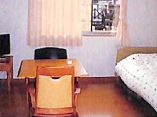 居室にはテーブルや椅子、ベッドが完備されている。テレビも設置されている。奥には窓があり、カーテンが掛けられている。