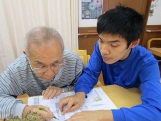介護スタッフと利用者の活動 デイサービス 利用者と並んで座り、丁寧に活動を支援しているスタッフがいます。温かいコミュニケーションを大切にしており、就業にやりがいを感じられる環境です。