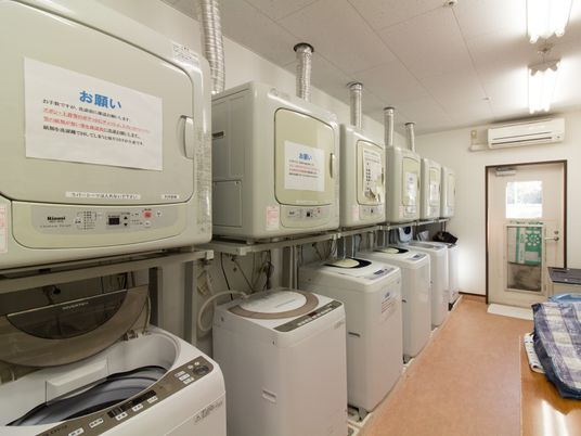 乾燥機付き全自動洗濯機が６台並んでいる。乾燥機の扉に注意書きが貼られている。エアコンが設置されている。