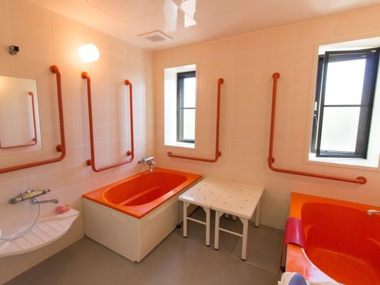 サポートの必要な入居者様用の浴室が用意されている。オレンジの浴槽が２つあり、壁際に腰掛が２つ置かれている。