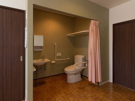 居室のトイレは、ドアではなくカーテンで仕切られている。天井近くに棚が１段あり、壁に手すりが設置されている。