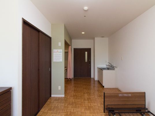居室の床は格子状の模様のフローリング材である。壁にカレンダーが下げられている。小型のキッチンが用意されている。