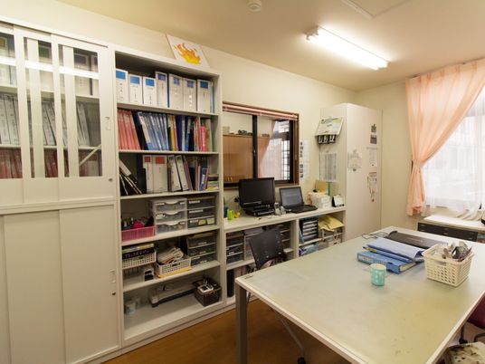 部屋の中央には大きな机があり、ファイルがいくつか置かれている。パソコンの前にある窓から、受付業務をすることができる。