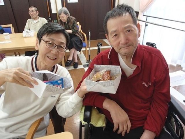 笑顔の利用者と職員 ご入居者様 職員と利用者が笑顔でおやつを楽しむ光景が、温かい雰囲気の食堂を示しています。利用者の方々の快適な日常を支えるお仕事です。