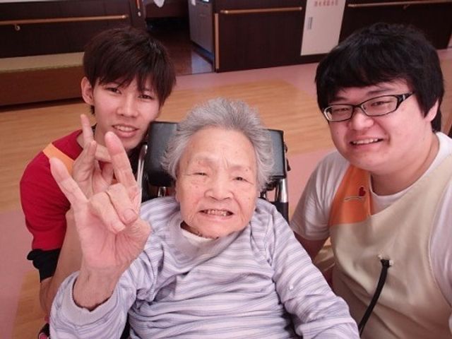 笑顔の方々と共有スペース スタッフとご入居者様 笑顔でポーズをとる利用者様とスタッフが印象的な共有スペースで、心温まる交流の一コマが垣間見えます。安心してご就業いただける雰囲気が広がっています。