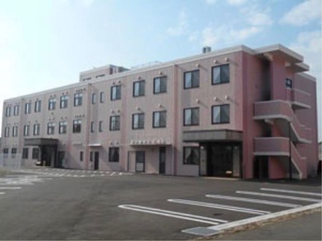 ピンクの建物の外観 施設外観 広々とした駐車場が整備されたモダンな設計の建物です。働く環境として、清潔感があり、快適さが感じられます。