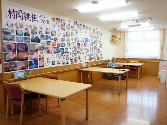 壁に写真多数の休憩室 談話コーナー 壁にはカラフルな写真がずらりと飾られ、ゆったりとした空間の休憩室で1人の方が静かに過ごされています。利用者の皆様の憩いの場で、ここで新しい仲間との出会いも期待できます。