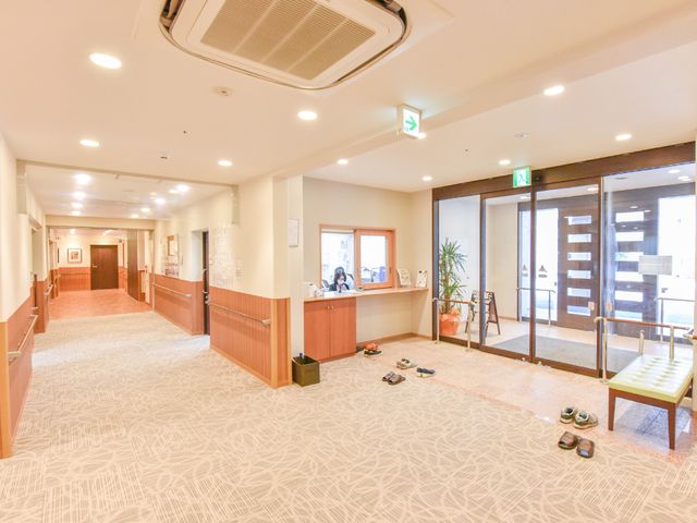 広々とした明るいロビー エントランス 落ち着いた色合いの広いロビーには、明るい照明があり、受付でスタッフが業務にあたっています。靴を脱いでいる様子から、訪れる方がリラックスして過ごせる雰囲気が伝わります。