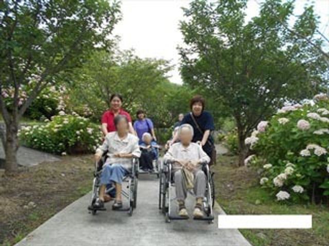車椅子で散歩する人々とスタッフ 外出イベント スタッフが優しく寄り添いながら、利用者様が爽やかな中庭の散策を楽しんでいらっしゃいます。充実した緑に囲まれた環境で、気持ちの良い時間を過ごせる場所です。