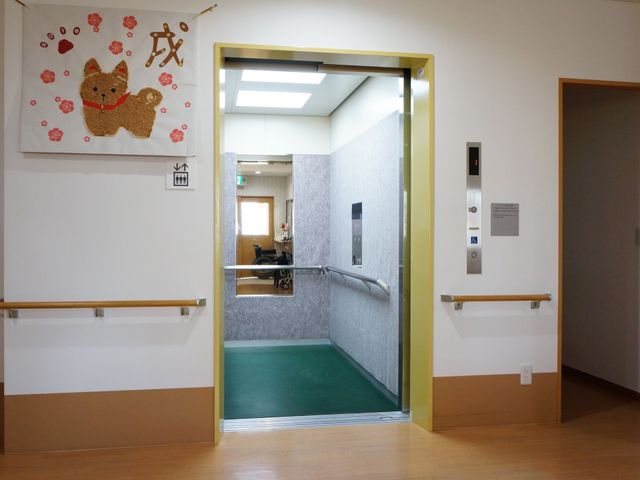 廊下とエレベーター エレベーター 手すりが設置された温かみのある廊下が見えます。車椅子が停められるスペースも確保され、ゆったりとした動線が実現されています。