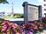 サムネイル 当施設名と事業内容が書かれた看板である。木材で覆われた看板の周りにはピンクや紫の明るい花々が色を添えている。