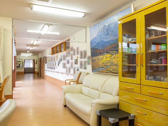 暖かみのある廊下 廊下 ゆったりとしたソファが設置され、休憩に適した明るく開放感のあるスペースです。壁には掲示物や絵画が飾られ、温かな雰囲気を演出しています。