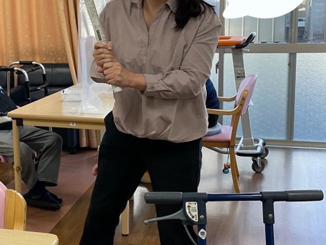 笑顔のスタッフと居室 スタッフイメージ 手にはさみを持ち、笑顔で立つスタッフが明るい居室にいます。周囲には椅子があり、居心地の良さが感じられます。