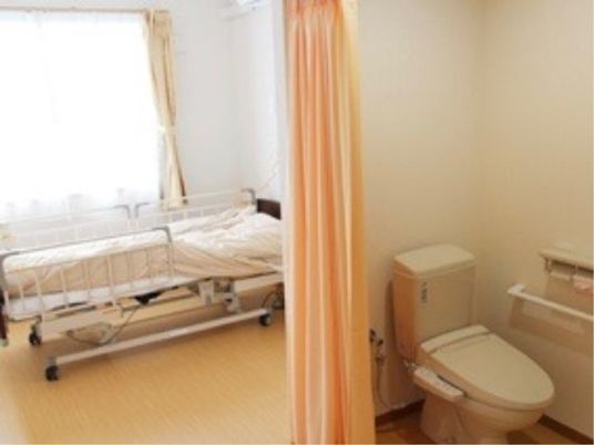 腰高窓の前には介護ベッドが置かれている。部屋の手前側に温水洗浄便座のついたトイレがある。便器の横には手すりが付いている。