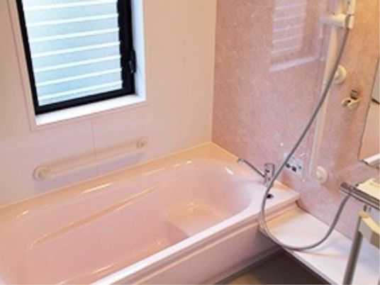 シンプルな形状の個浴である。窓から自然光が差し込んでいる。浴槽の向こう側の壁と手間の壁に手すりが付いている。