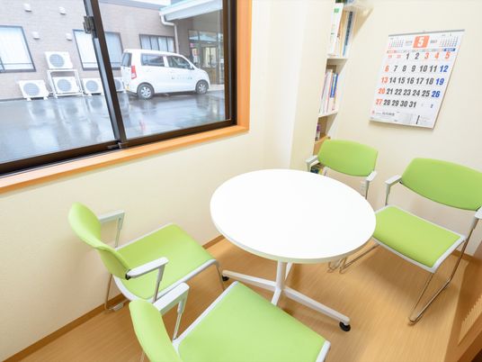 白い円卓を囲んで、緑色の椅子が４脚置かれている。壁にはカレンダーと本棚が取り付けられており、外には施設専用車が停められている。