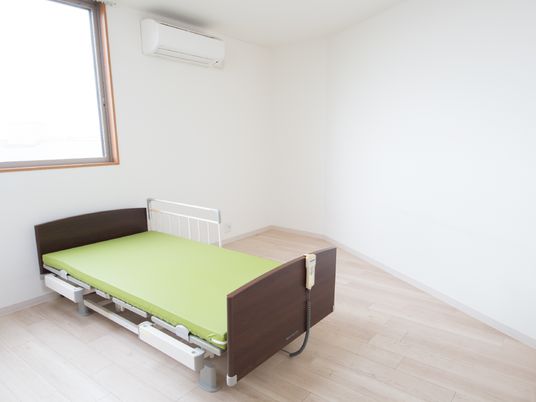 角部屋は、壁が斜めになった部分がある。大きな窓の下に介護用ベッドがあり、天井近くにエアコンが設置されている。