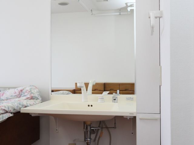 居室の洗面台とベッド 個室の洗面台 ゆとりのある居室を持つ施設で、機能的な洗面台の様子です。快適な毎日をサポートする設備が整っています。