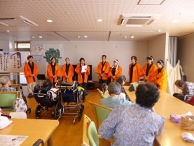 集合するスタッフと利用者 リビング 明るい多目的室にて、スタッフが集まり何かの説明をしている様子です。利用者の方々が集い、和やかな交流の場が広がっています。