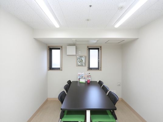 施設の写真 縦長の窓が２つある室内である。黒いテーブルとグリーンの椅子が６脚設置されていて、テーブルにはパンフレットなどがのっている。