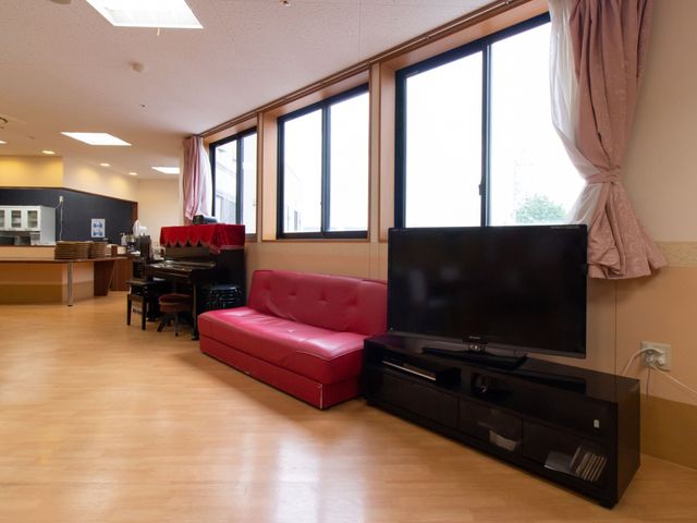 共有スペースの様子 共有スペース 広々としたラウンジには、明るい日差しが差し込み、大型テレビを設置したリラックスできる空間が広がっています。赤のソファが印象的で憩いのひとときを提供しています。