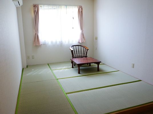 床には畳が敷いてある。奥には窓があり、その手前には椅子とテーブルがそれぞれ1つ置かれている。左上奥にはエアコンが付いている。