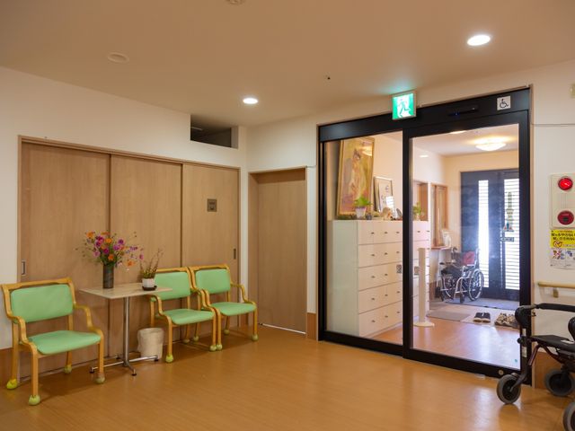 待合室の椅子と受付 出入口 清潔感あふれる待合室には、優しい色合いの椅子が並び、左手に見える受付は来訪者の対応に備えています。壁にはアートが飾られ、窓越しには車椅子を利用されている方が視認でき、落ち着いた雰囲気が伝わってきます。