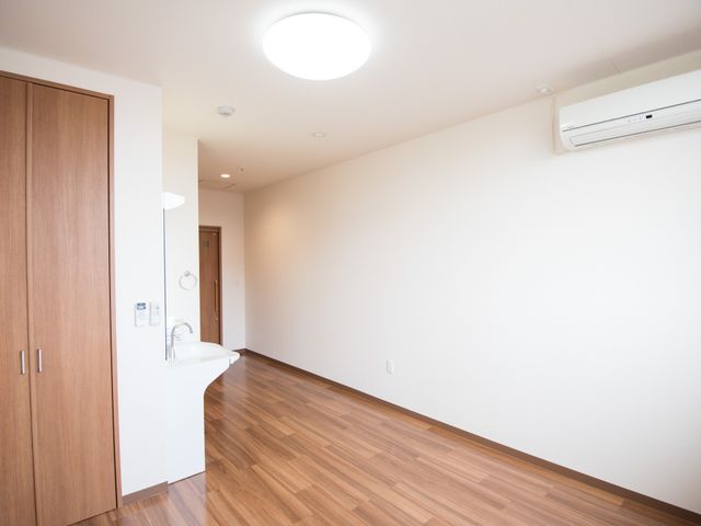居室の入口と洗面台 居室 シンプルで暖かみのある床材の居室です。清潔感溢れる洗面台が設置され、快適な生活空間が保たれています。