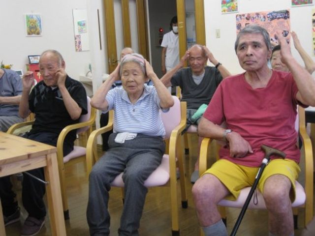 機能訓練室での活動 入居者様 明るい雰囲気の中で、スタッフのサポートのもと、体操を行っている様子がうかがえます。参加者は元気に活動しており、健康維持に励んでいるようです。