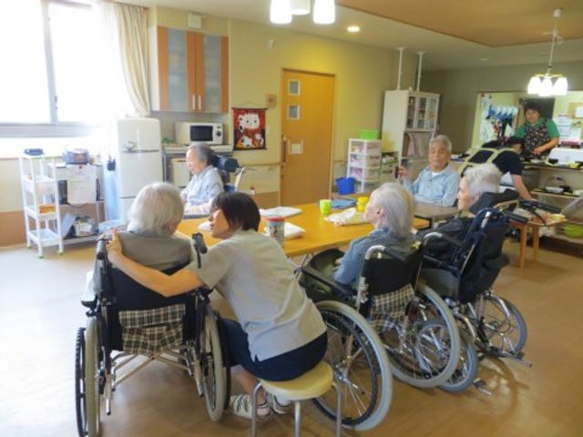 車椅子の利用者とスタッフ リビング スタッフが利用者の方に寄り添い、明るい食事スペースでコミュニケーションが図られています。車椅子利用者も安心して過ごせる設計です。