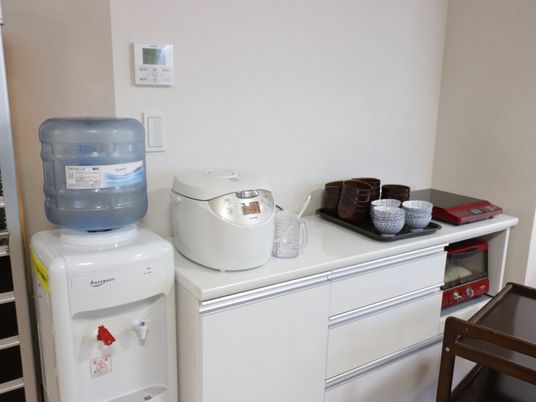 食器棚の上に炊飯器、電磁調理器、湯飲みが置かれている。隣にはウォーターサーバーも備わっており、自由に水を飲むことができる。