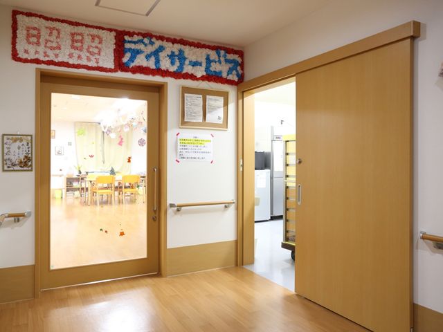 快適な食堂の入口 デイサービス室 床がきれいで落ち着いた色合いの入口から見える食堂は、開放的な雰囲気でご利用者が集う場所です。窓から自然光が入り、明るく温かみのある空間をご覧いただけます。